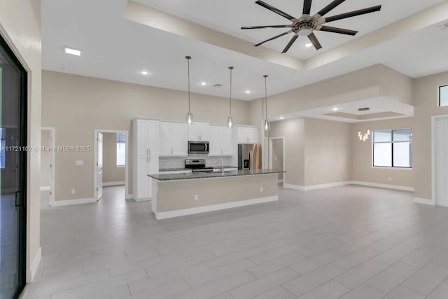 kitchen featuring a kitchen island with sink, stainless steel appliances, a tray ceiling, white cabinets, and decorative light fixtures