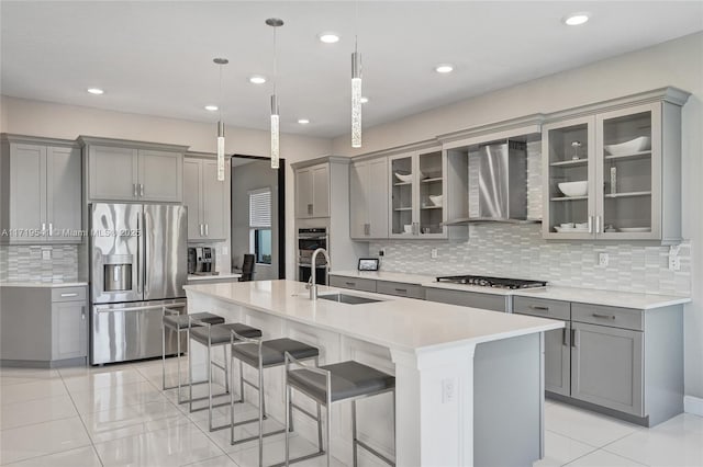 kitchen with sink, wall chimney exhaust hood, stainless steel appliances, decorative light fixtures, and a center island with sink