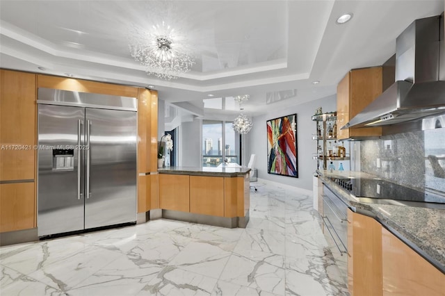 kitchen featuring wall chimney exhaust hood, built in refrigerator, an inviting chandelier, and a raised ceiling