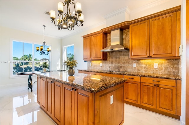 kitchen featuring marble finish floor, hanging light fixtures, brown cabinetry, dark stone counters, and wall chimney exhaust hood