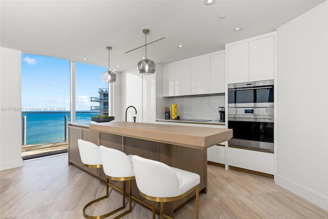 kitchen featuring multiple ovens, an island with sink, decorative light fixtures, a water view, and white cabinets