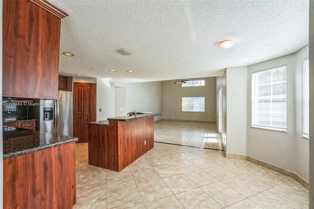 kitchen featuring dark stone counters, stainless steel fridge with ice dispenser, a center island with sink, and ceiling fan