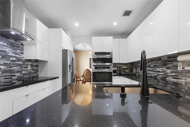 kitchen with white cabinets, wall chimney exhaust hood, stainless steel appliances, and dark stone counters