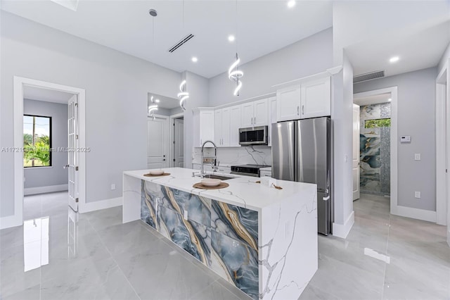 kitchen with a center island with sink, hanging light fixtures, light stone countertops, white cabinetry, and stainless steel appliances