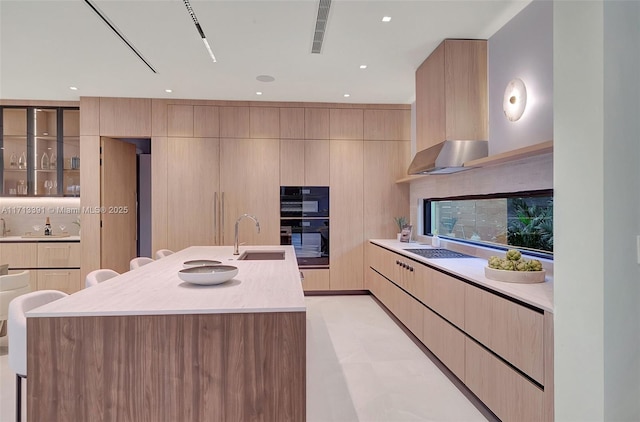 kitchen with dobule oven black, a kitchen island with sink, light countertops, wall chimney range hood, and a sink