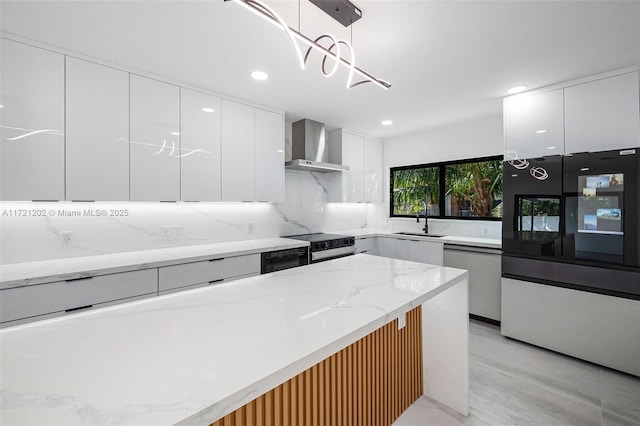 kitchen featuring white cabinets, hanging light fixtures, stainless steel dishwasher, light stone countertops, and wall chimney range hood