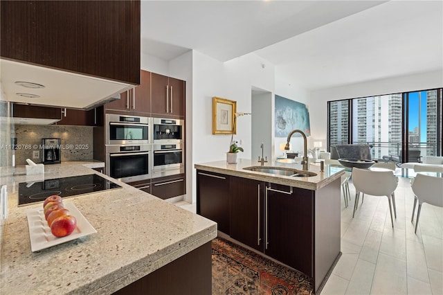 kitchen featuring an island with sink, black electric cooktop, tasteful backsplash, light stone counters, and sink