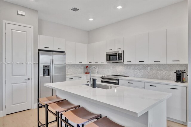 kitchen with sink, a center island with sink, appliances with stainless steel finishes, a kitchen breakfast bar, and white cabinets