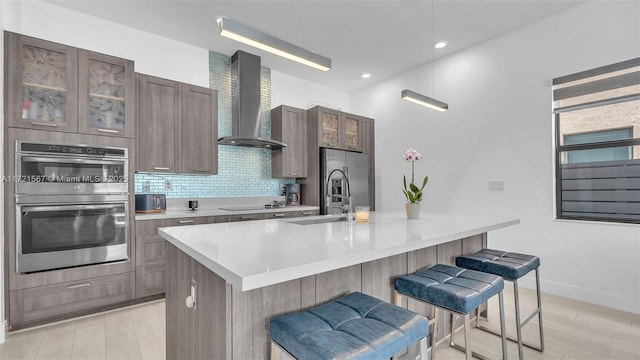 kitchen with a breakfast bar, a center island with sink, wall chimney range hood, sink, and stainless steel appliances