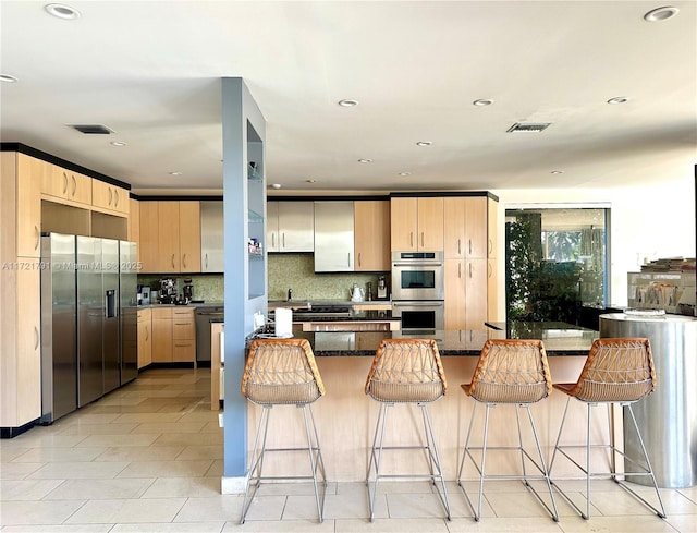 kitchen with stainless steel appliances, dark stone counters, decorative backsplash, light brown cabinetry, and light tile patterned floors