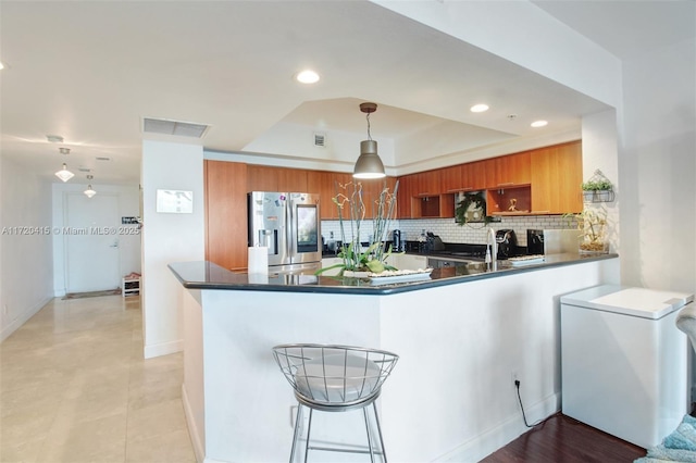 kitchen with kitchen peninsula, stainless steel refrigerator with ice dispenser, a breakfast bar, a raised ceiling, and pendant lighting