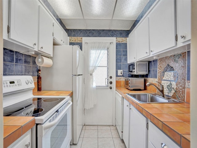 kitchen with a paneled ceiling, white appliances, sink, tile countertops, and white cabinets