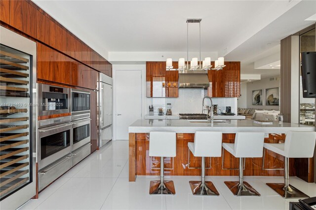 kitchen with hanging light fixtures, stainless steel built in refrigerator, light tile patterned flooring, a kitchen bar, and wall chimney range hood