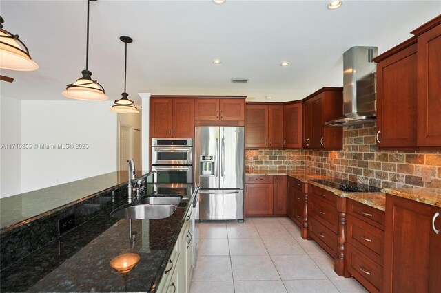 kitchen featuring decorative light fixtures, dark stone counters, wall chimney range hood, stainless steel appliances, and a center island with sink