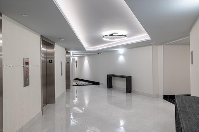 hallway featuring light tile patterned floors, a tray ceiling, elevator, and baseboards
