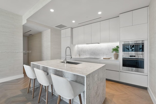 kitchen with sink, stainless steel double oven, a kitchen island with sink, light parquet floors, and white cabinets