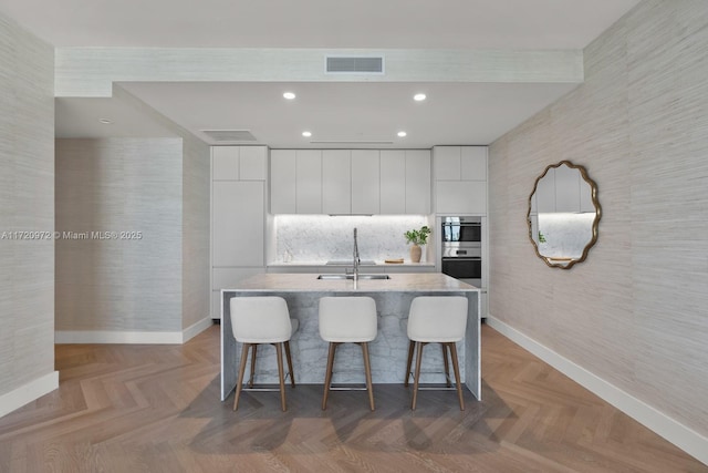 kitchen with parquet flooring, sink, white cabinetry, an island with sink, and oven