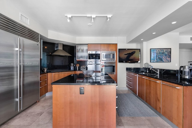 kitchen featuring track lighting, sink, wall chimney range hood, built in appliances, and a center island