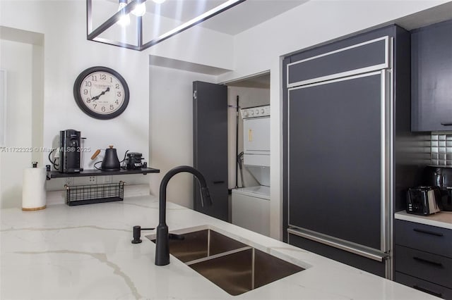 kitchen with paneled built in refrigerator, light stone counters, stacked washer / dryer, and sink