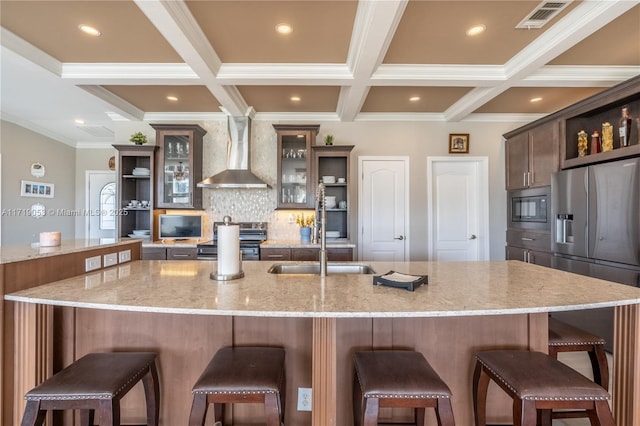 kitchen with wall chimney range hood, a kitchen breakfast bar, light stone counters, a large island with sink, and appliances with stainless steel finishes