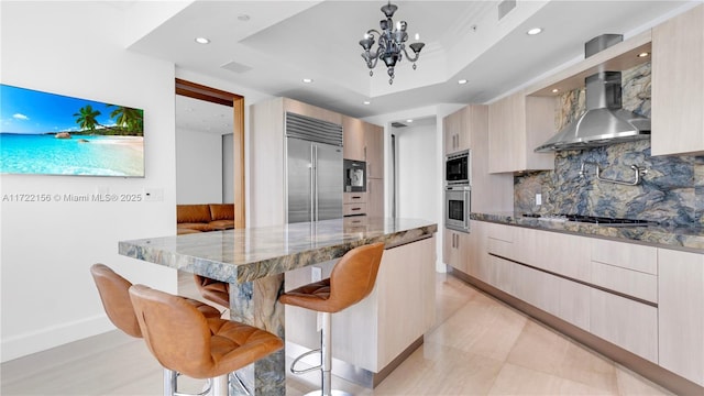 kitchen featuring a breakfast bar, a raised ceiling, wall chimney range hood, built in appliances, and light stone countertops