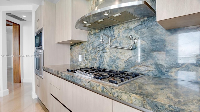 kitchen with light brown cabinets, range hood, backsplash, stone countertops, and appliances with stainless steel finishes