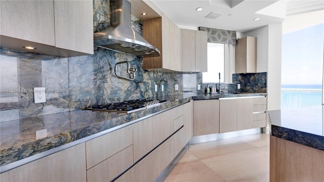 kitchen with wall chimney range hood, tasteful backsplash, dark stone counters, stainless steel gas stovetop, and light brown cabinetry