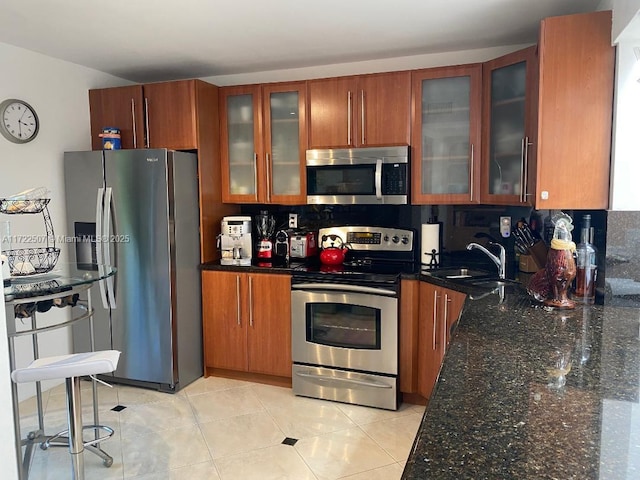 kitchen with appliances with stainless steel finishes, a sink, brown cabinetry, dark stone countertops, and backsplash