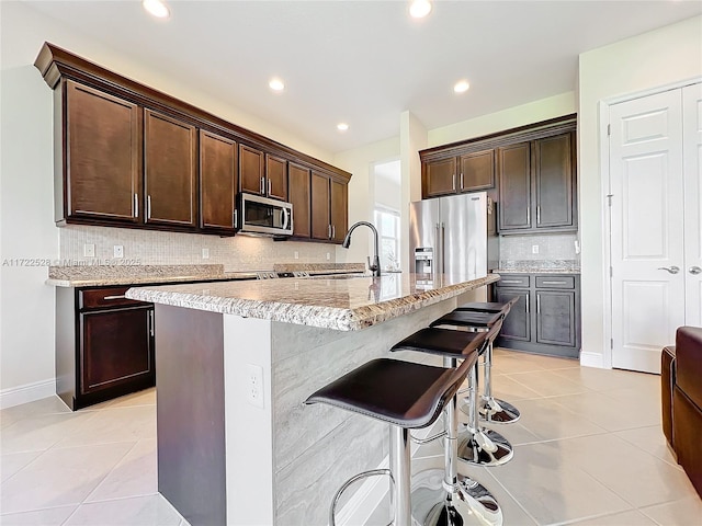 kitchen featuring a kitchen island with sink, sink, light tile patterned floors, appliances with stainless steel finishes, and dark brown cabinets