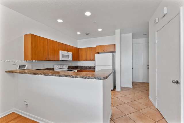 kitchen with white appliances, kitchen peninsula, light tile patterned floors, and dark stone counters