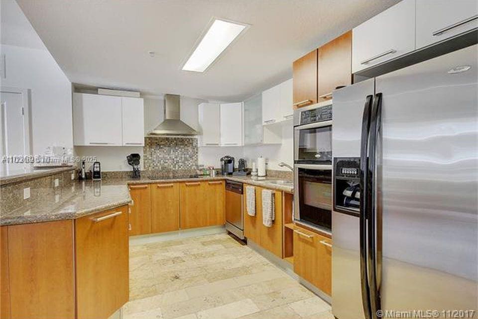 kitchen featuring wall chimney exhaust hood, stone countertops, white cabinetry, and appliances with stainless steel finishes