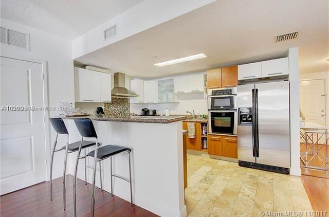 kitchen with a kitchen bar, kitchen peninsula, stainless steel appliances, wall chimney range hood, and white cabinets