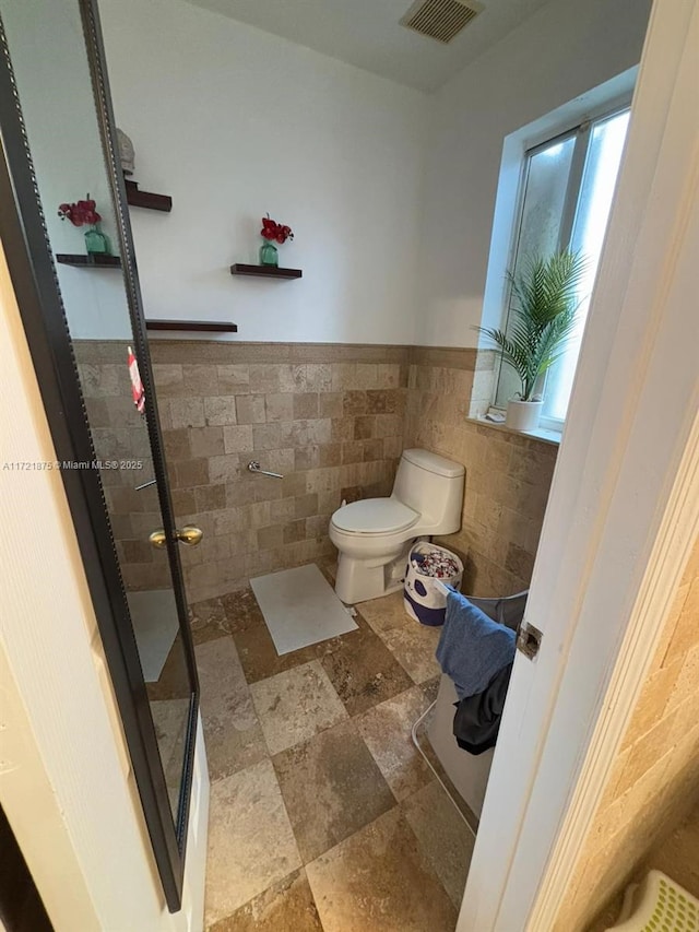 bathroom with visible vents, toilet, a wainscoted wall, walk in shower, and tile walls