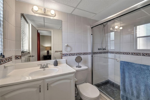 bathroom featuring a drop ceiling, vanity, a shower with door, toilet, and tile walls