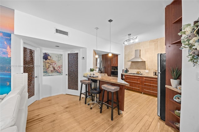 kitchen with decorative backsplash, a kitchen breakfast bar, stainless steel appliances, wall chimney range hood, and decorative light fixtures