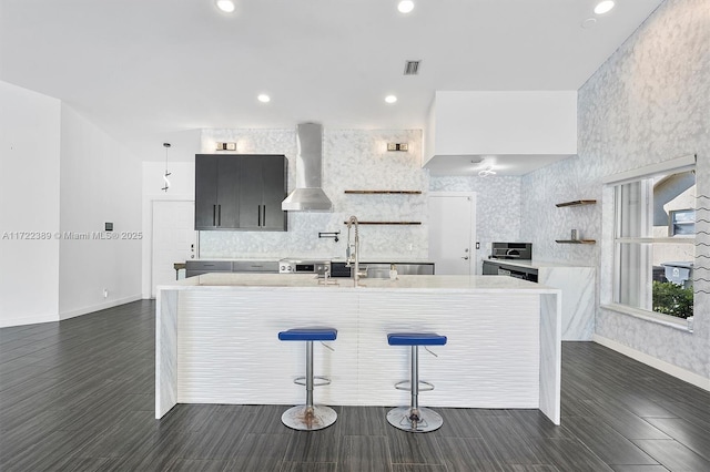 kitchen featuring sink, wall chimney range hood, tasteful backsplash, a breakfast bar, and a center island with sink