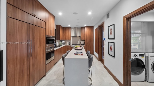 kitchen with a kitchen breakfast bar, wall chimney exhaust hood, light tile patterned floors, washing machine and clothes dryer, and a kitchen island