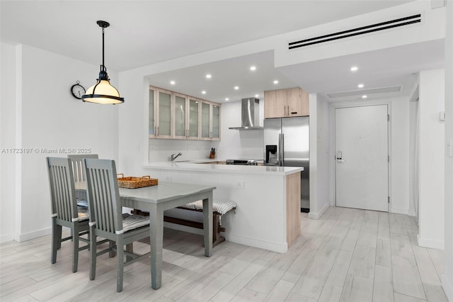 kitchen featuring kitchen peninsula, stainless steel refrigerator with ice dispenser, light wood-type flooring, wall chimney range hood, and light brown cabinets