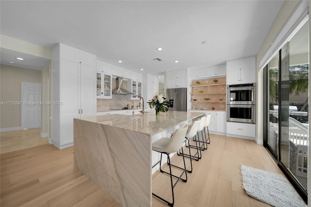 kitchen featuring appliances with stainless steel finishes, a large island with sink, white cabinetry, and wall chimney range hood