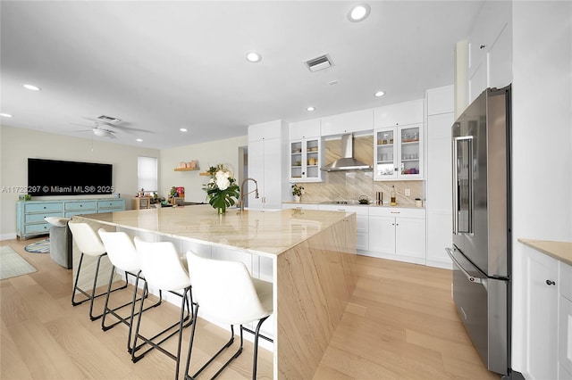 kitchen featuring a spacious island, high end refrigerator, wall chimney range hood, light stone counters, and a breakfast bar