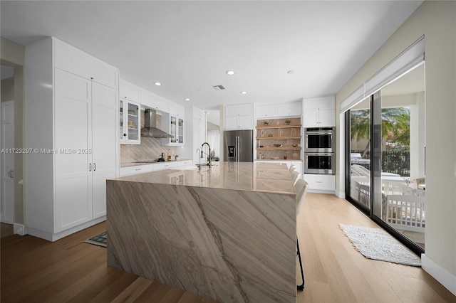 kitchen featuring white cabinets, wall chimney range hood, appliances with stainless steel finishes, and a large island with sink