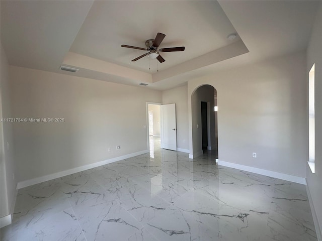 unfurnished room featuring arched walkways, a raised ceiling, baseboards, a ceiling fan, and light marble finish flooring