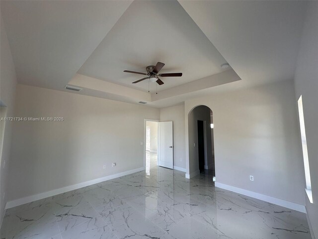 empty room with arched walkways, a tray ceiling, ceiling fan, baseboards, and light marble finish flooring