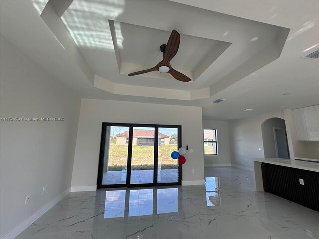 empty room featuring a tray ceiling, arched walkways, light marble finish floors, ceiling fan, and baseboards