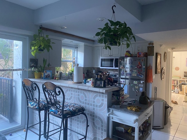 kitchen with tile countertops, white cabinetry, stainless steel fridge, a kitchen bar, and kitchen peninsula