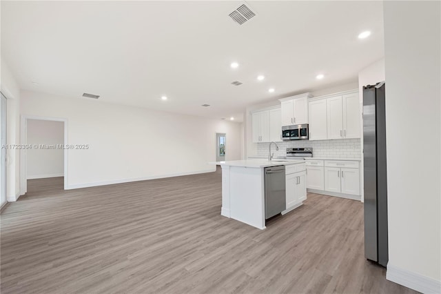 kitchen with light wood-type flooring, backsplash, stainless steel appliances, white cabinets, and an island with sink