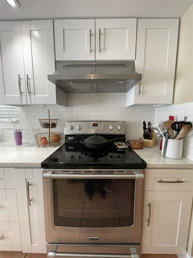 kitchen featuring backsplash, light stone counters, stainless steel range with electric stovetop, wall chimney range hood, and white cabinets