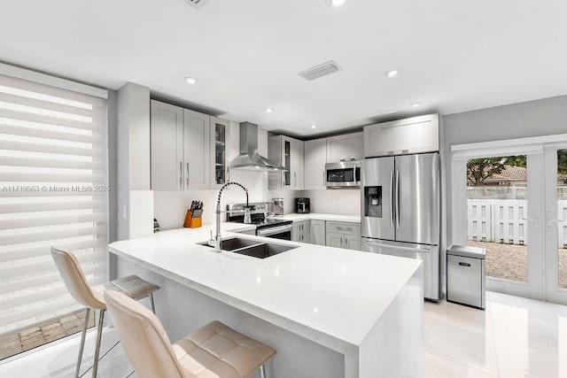 kitchen with kitchen peninsula, a kitchen breakfast bar, stainless steel appliances, sink, and wall chimney range hood