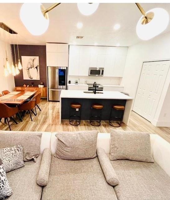 kitchen with appliances with stainless steel finishes, an island with sink, pendant lighting, white cabinets, and light wood-type flooring