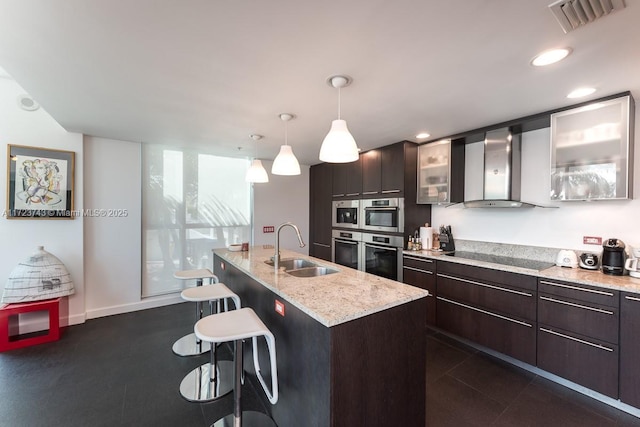 kitchen with wall chimney range hood, sink, black electric cooktop, an island with sink, and decorative light fixtures